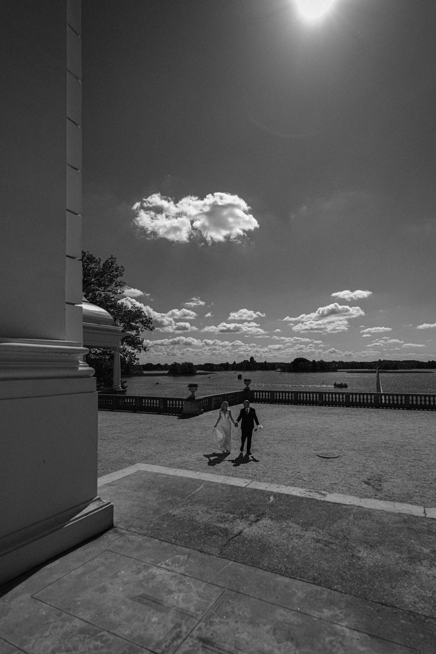 Black and white photo of a person walking on a terrace overlooking a river under a dramatic sky with a lone cloud.
