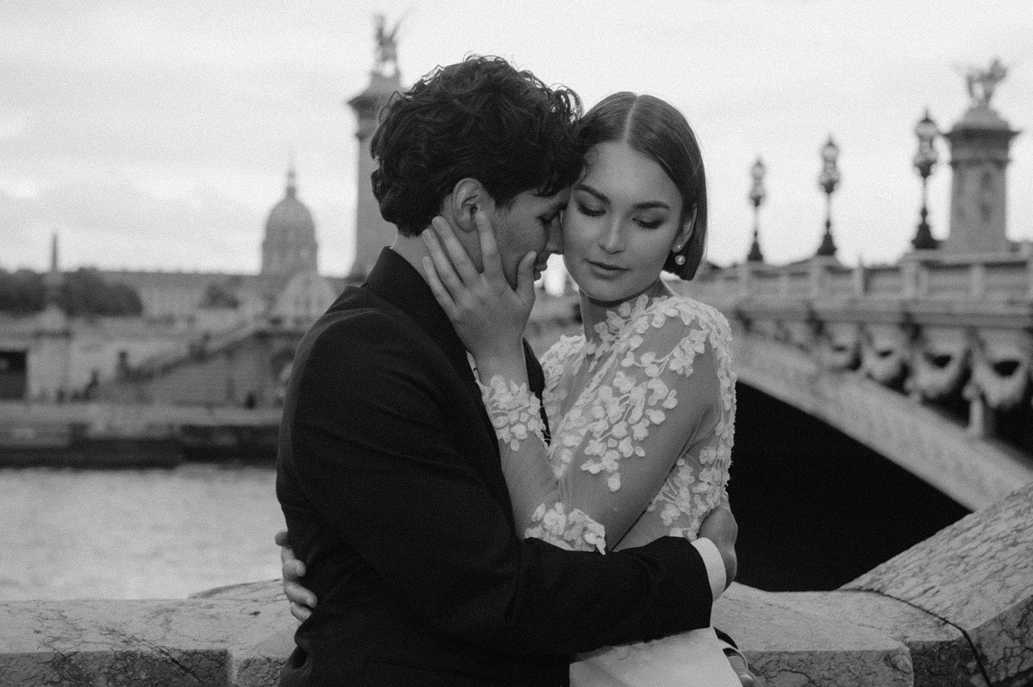 A romantic black and white photograph of a couple embracing on a bridge in Paris with ornate architecture in the background.