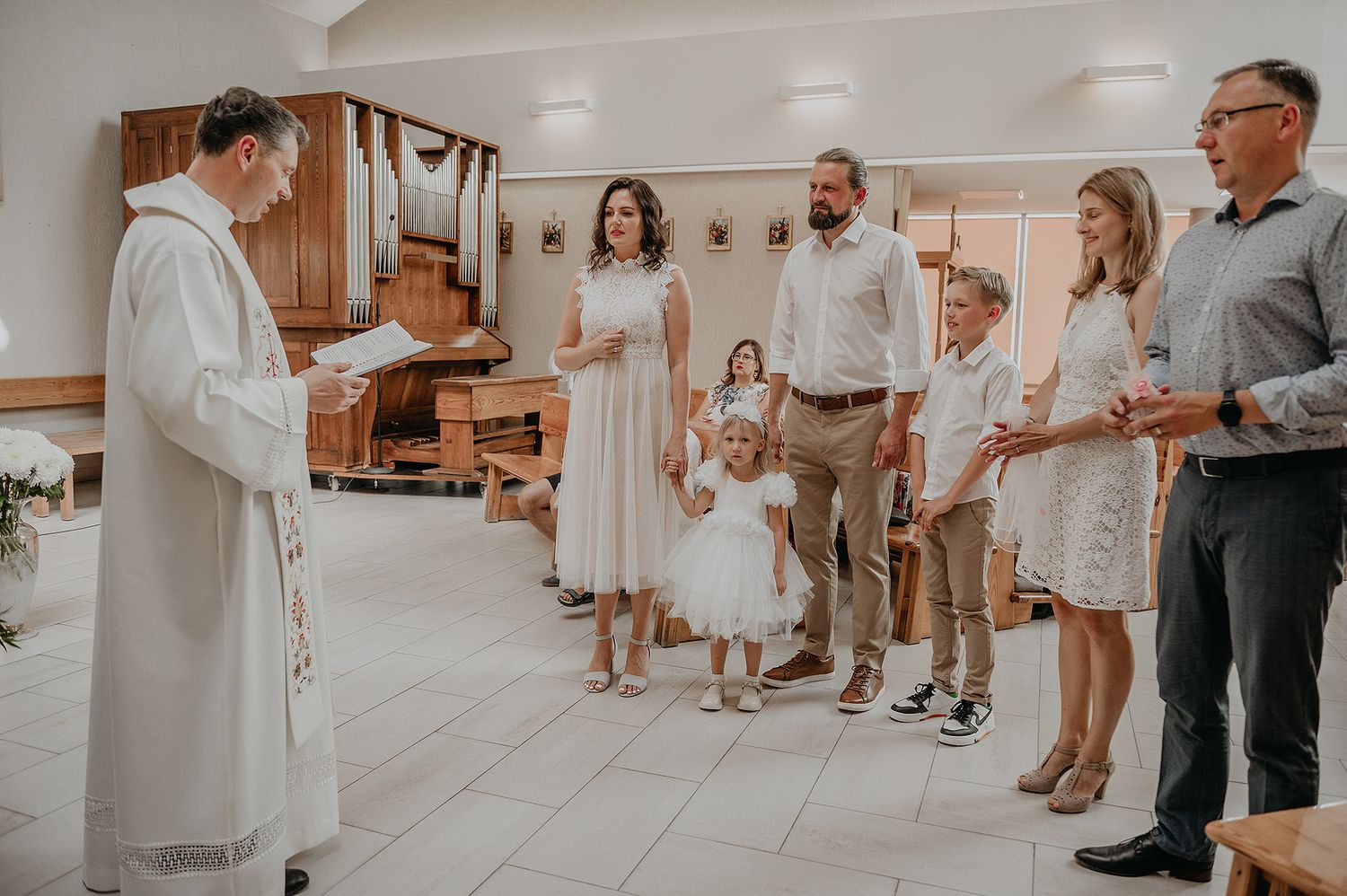 Wedding guests in light colored dresses and formal wear gather in a bright indoor space.