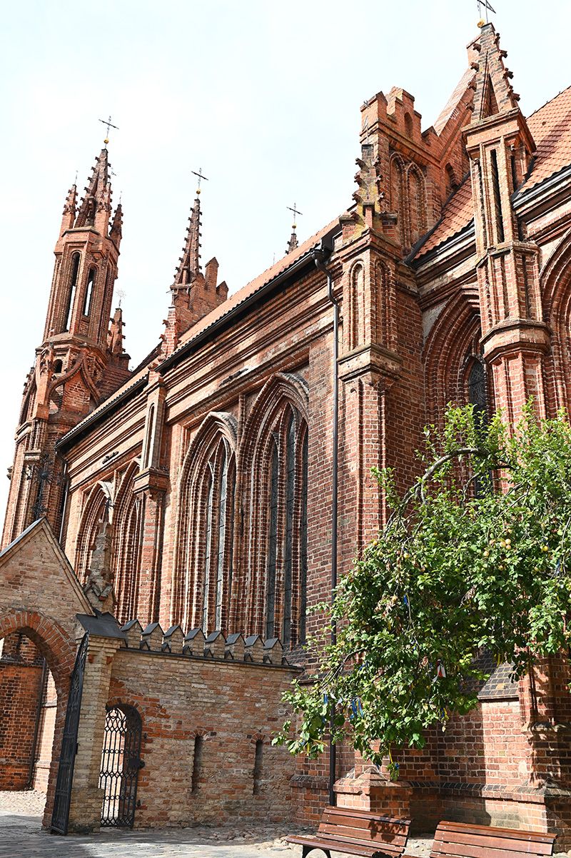 Gothic-style brick church with ornate spires and arched windows features intricate architectural details and green foliage.