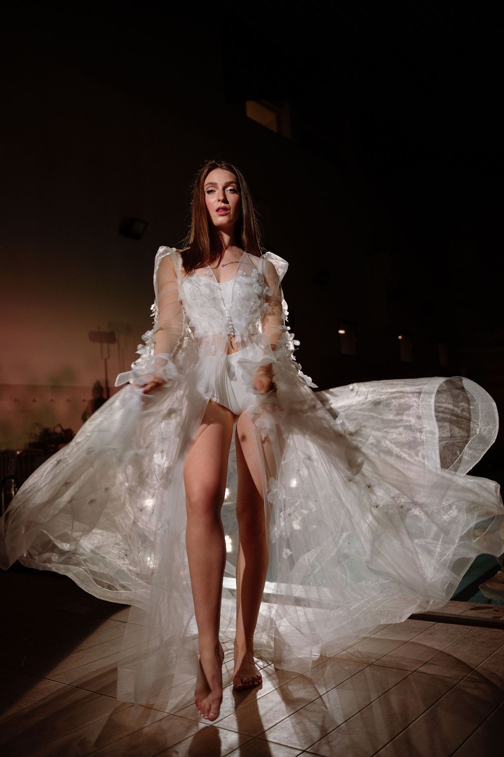 Model in a flowing sheer white gown with floral details walking barefoot on a wooden floor in dramatic low light.