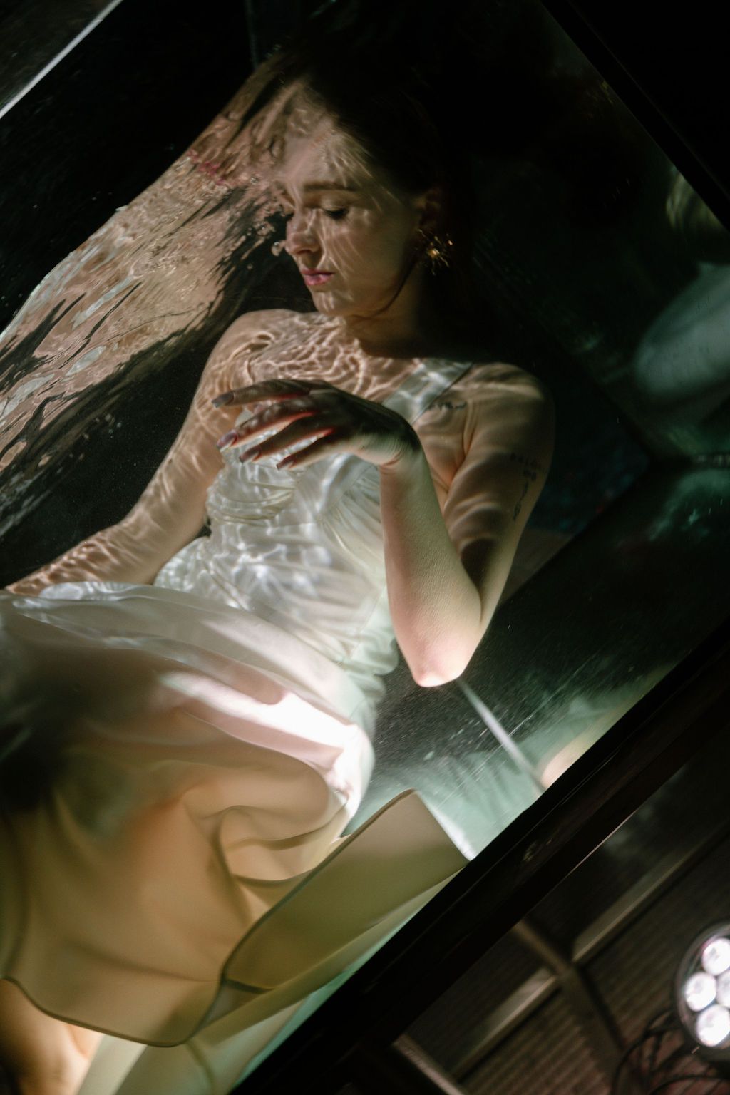 Woman in a flowing white dress reclines on a glass surface as rippling water light dances across her face and arms.