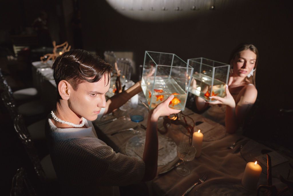 Two people at a candlelit table hold small glass tanks with goldfish, one wearing a pearl necklace, in a moody, dimly lit setting.