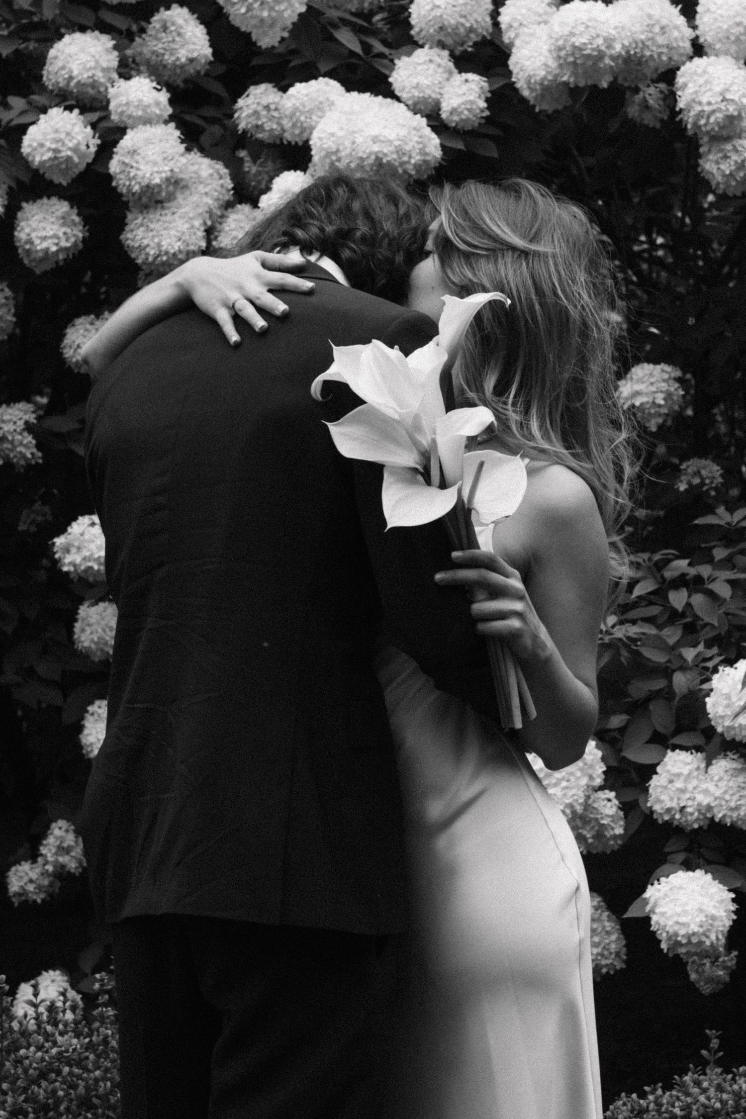 Black and white photo of a bride and groom embracing in a garden, she holds a calla lily bouquet against white hydrangea blooms.
