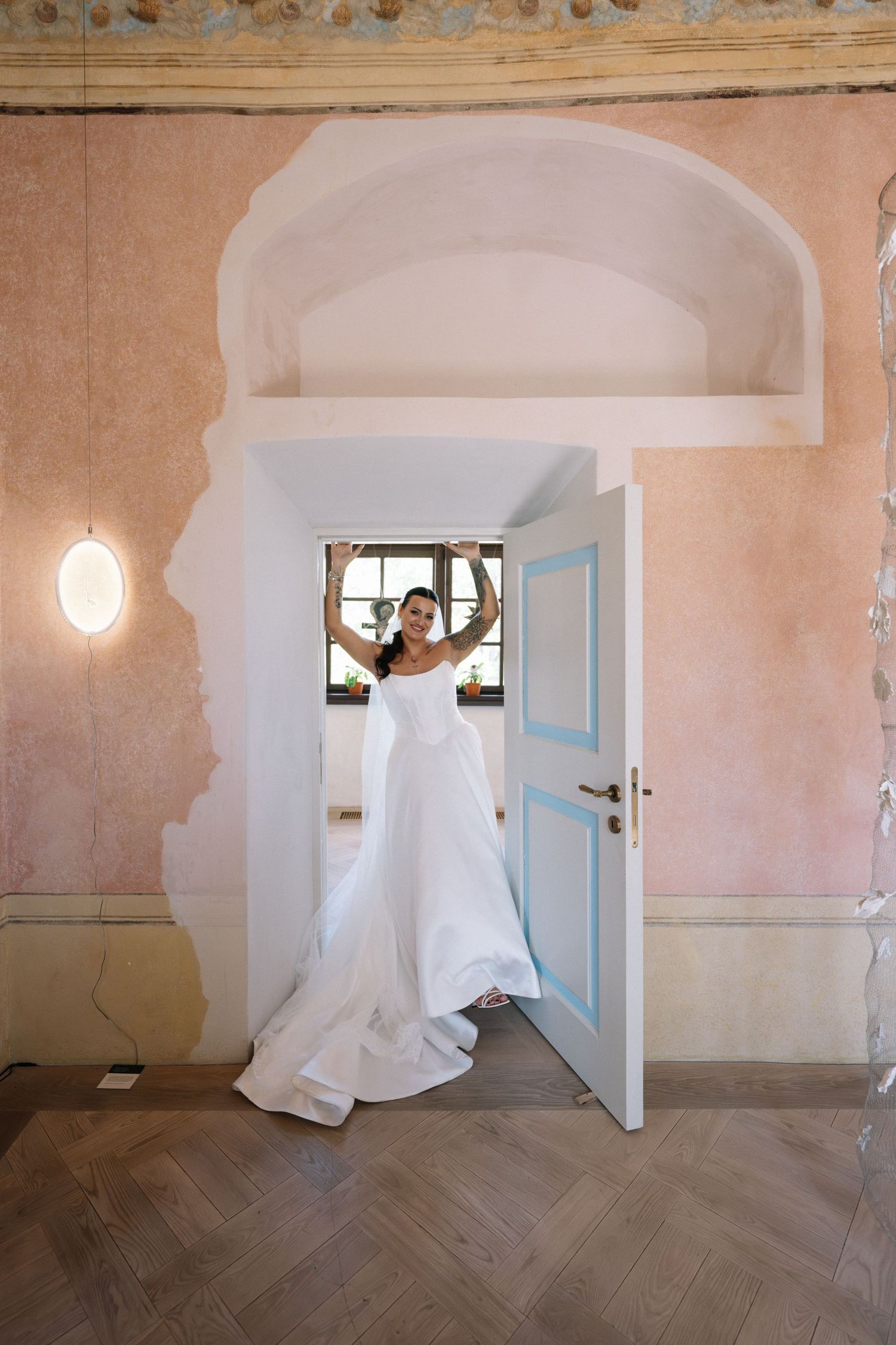 Bride in a flowing white gown raises her arms in an open blue-paneled doorway of a historic room with pink walls and herringbone floors.