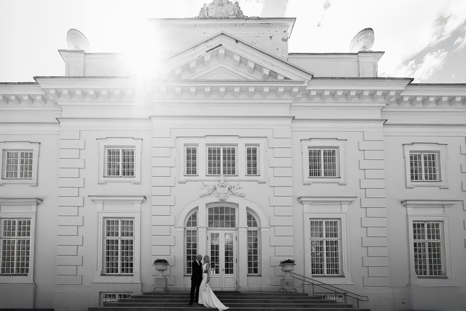 Bride in a white gown and groom in a dark suit pose on the steps of a grand neoclassical mansion in a black and white wedding photo.
