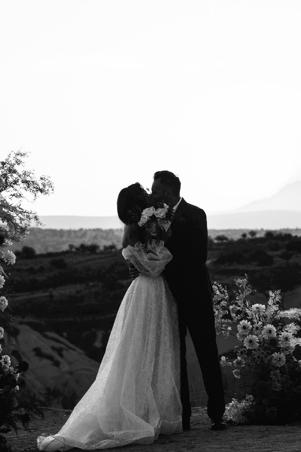 Black and white photo of a bride and groom sharing a kiss outdoors, with a scenic landscape and floral arrangements in the background.