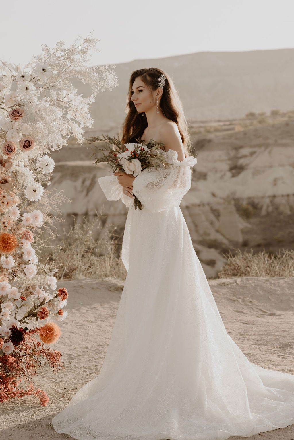 Bride in off-shoulder white gown holds bouquet by floral arch with desert mountains backdrop.