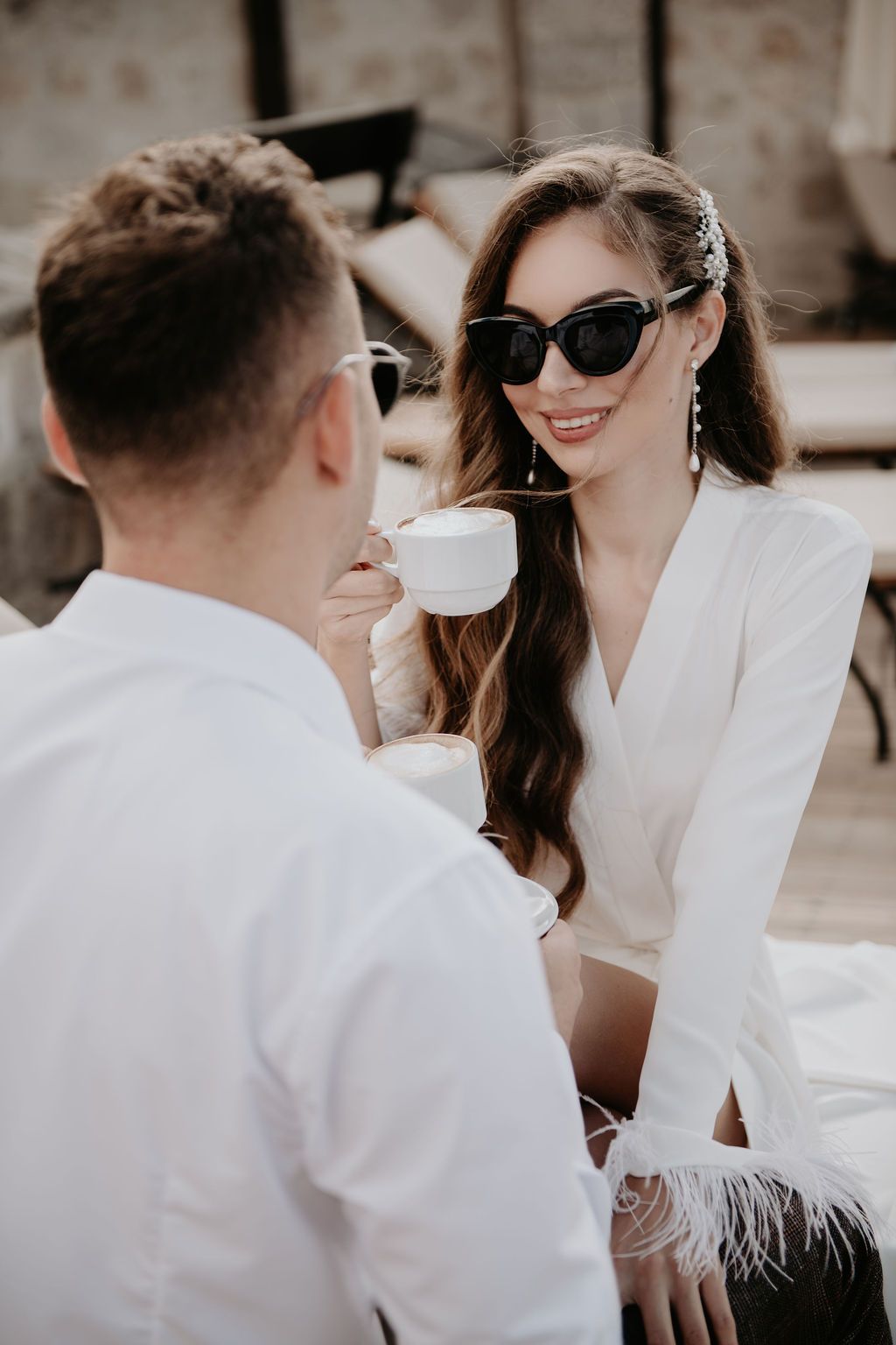 Elegant woman in white dress and sunglasses enjoying coffee with a man in white shirt at an outdoor cafe.