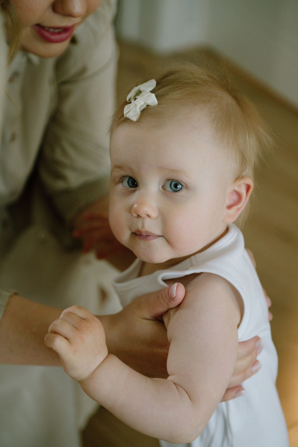 Adorable baby girl with blue eyes and white bow sits in white romper, supported by adult hands.