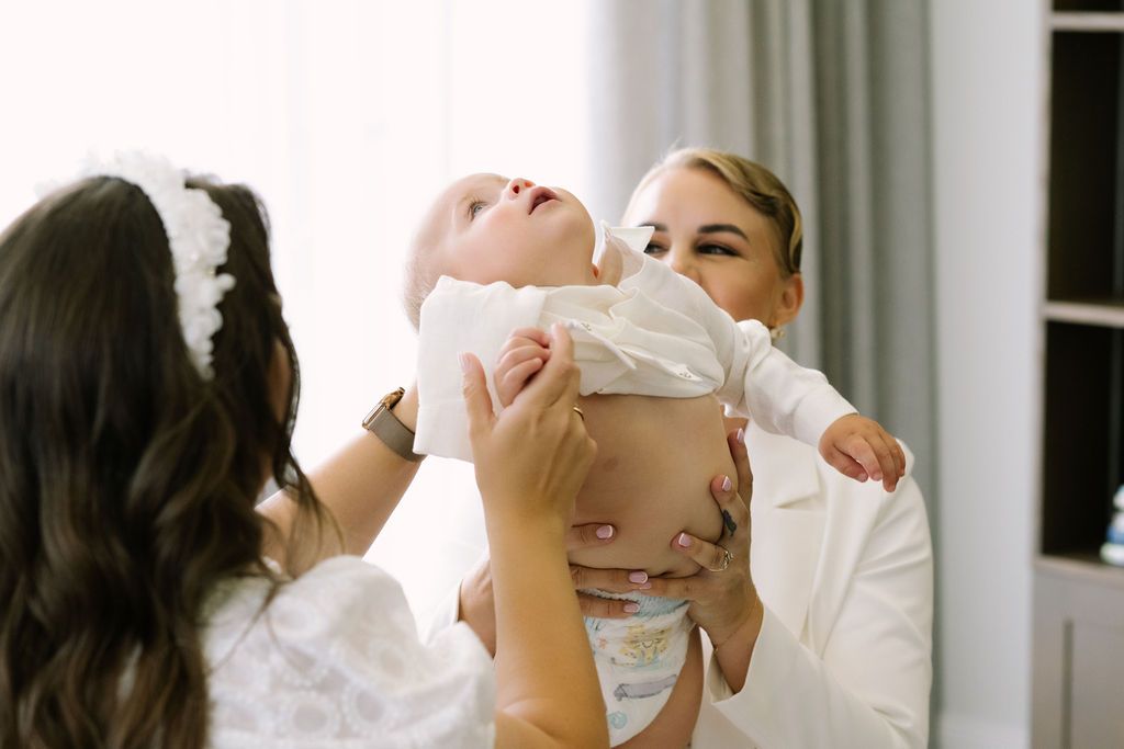 Woman in white holding baby up while another woman in white headband watches.