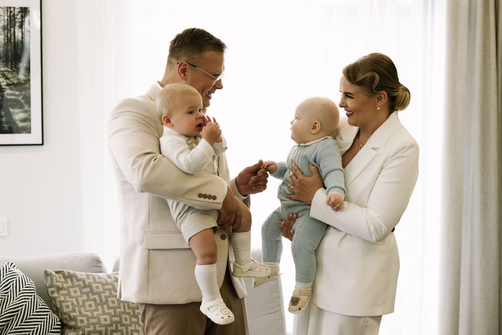 A couple in white suits smiling and holding two babies facing each other in a bright modern living room.