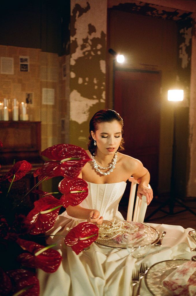 Elegant bride in white corset gown seated at decorated table with red anthurium flowers and pearl necklace in moody setting.