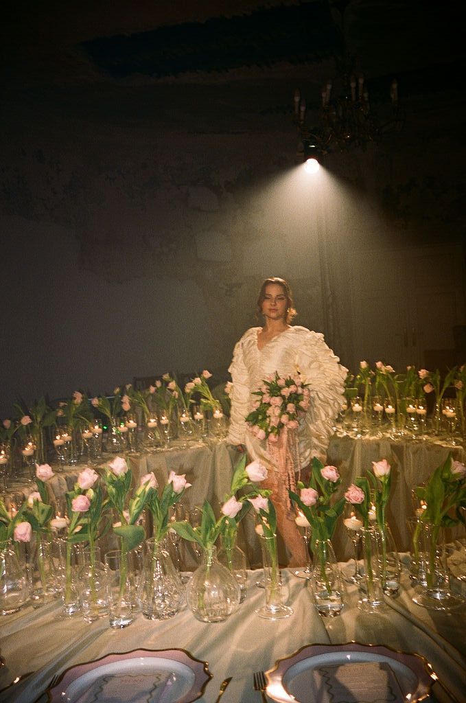 Woman in white ruffled gown holding pink bouquet surrounded by tulips in glass vases at elegant floral table setting.
