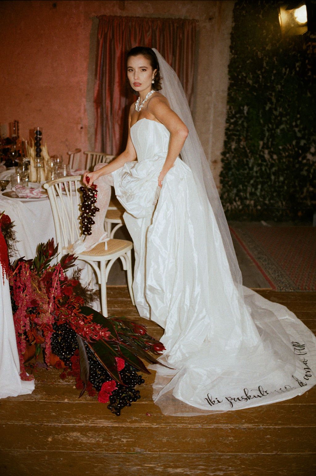Bride in strapless white ball gown with veil and pearl necklace poses beside red floral arrangements.