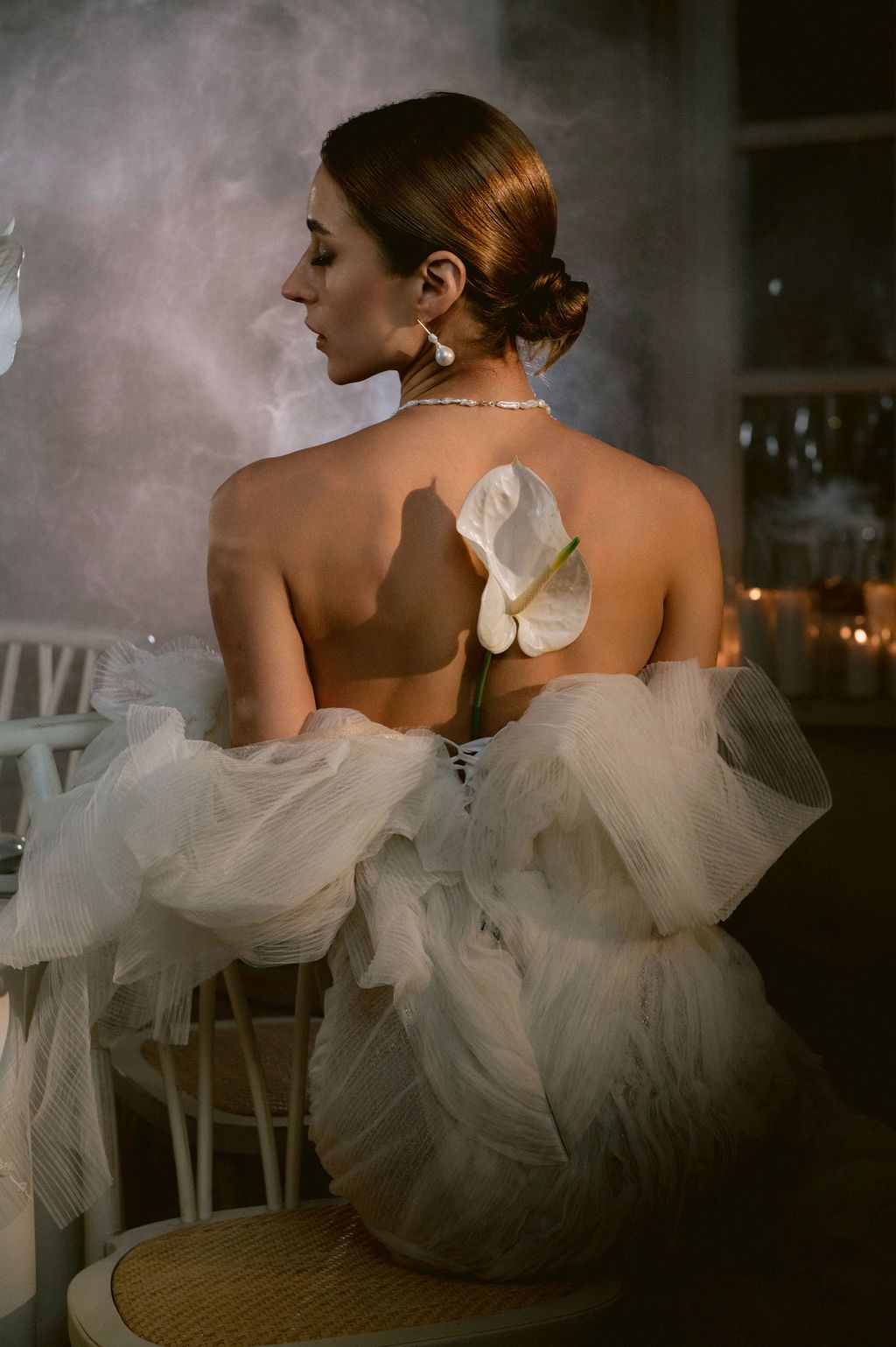 Elegant bride in backless white tulle gown with calla lily, pearl necklace, updo hairstyle, seated at candlelit reception.