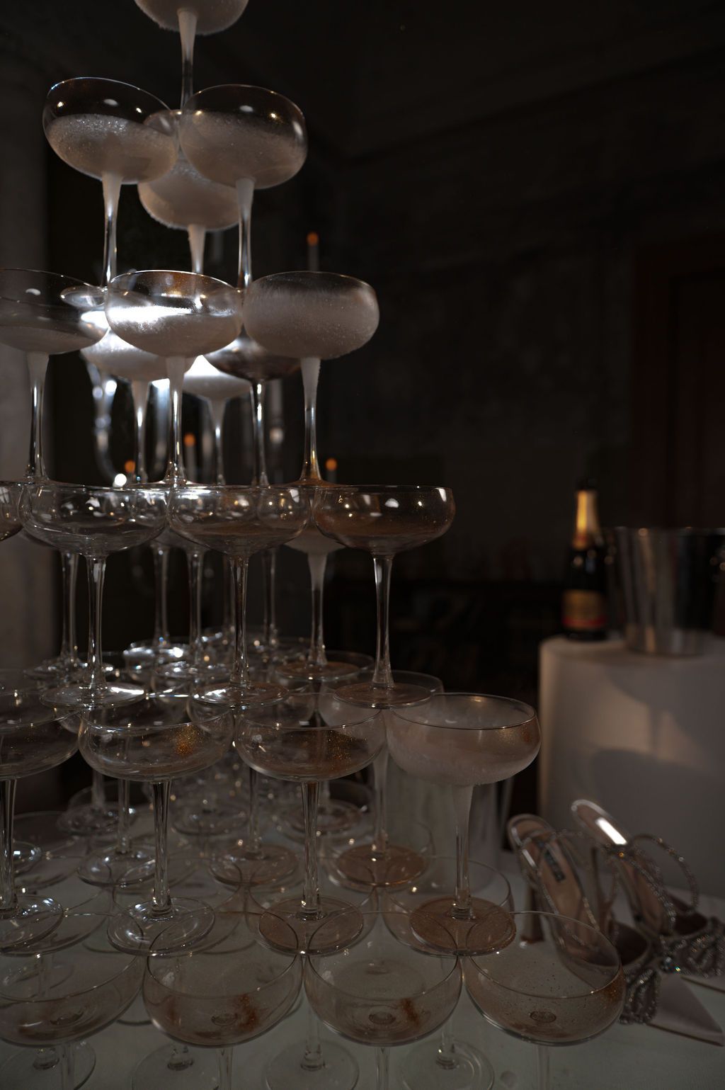 Multi-tiered champagne tower of coupe glasses at upscale celebration with bottle visible in background.