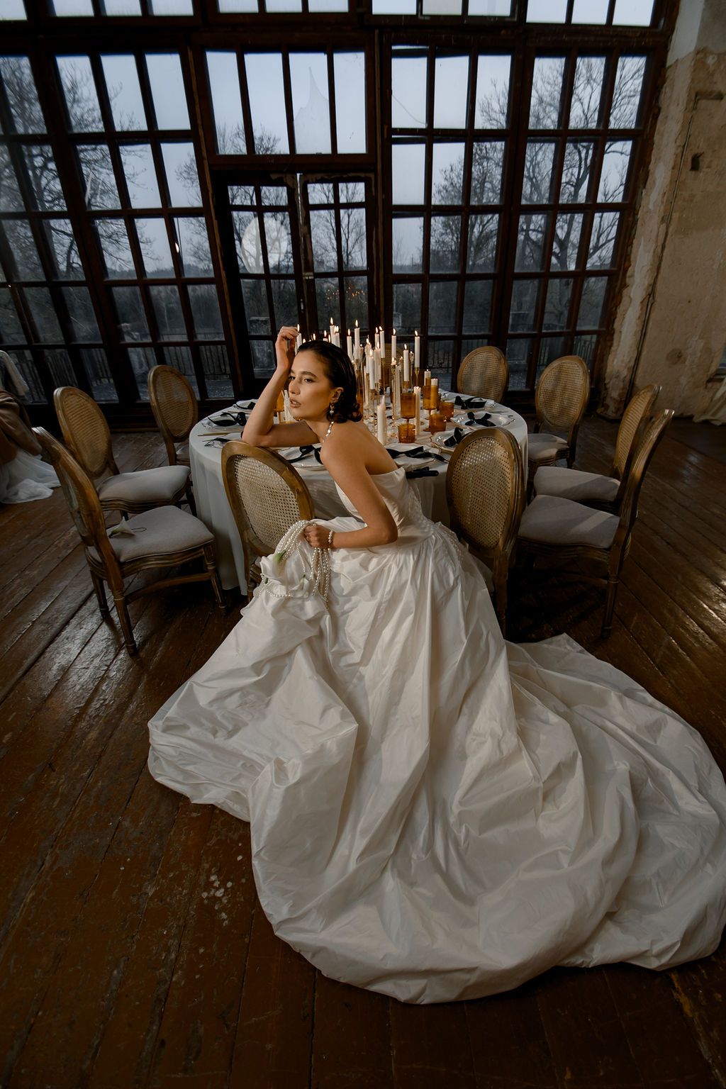 Bride in a voluminous white ball gown sits on hardwood floor beside candlelit reception table in elegant venue.