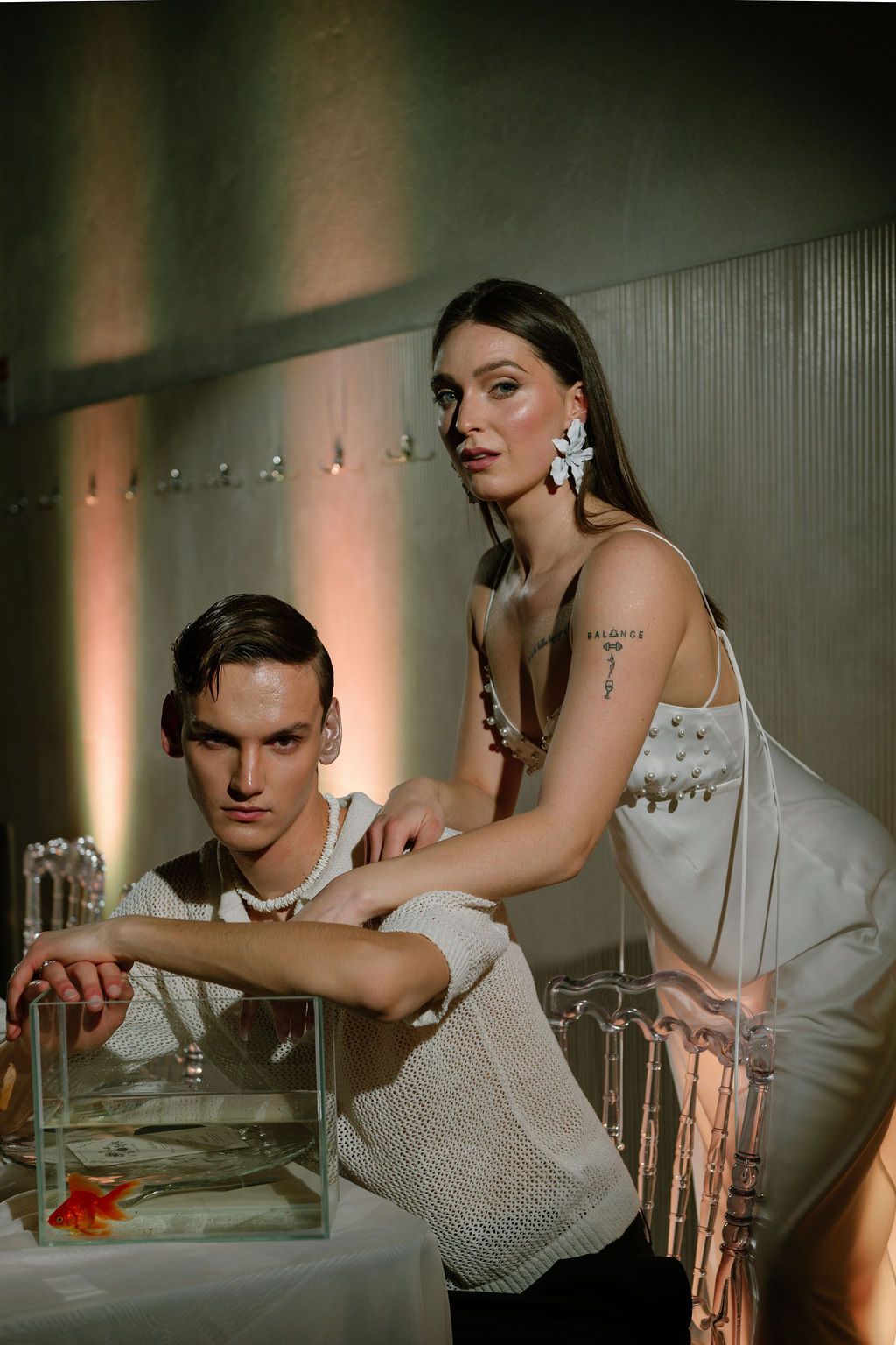 A man and woman pose elegantly beside a glass tank with a goldfish in a softly lit modern interior.