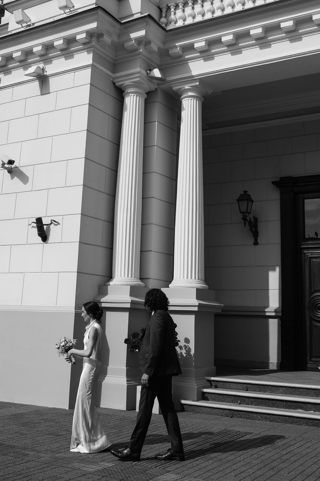 Black and white photo of a bride and groom walking in front of a classical building with tall white columns.