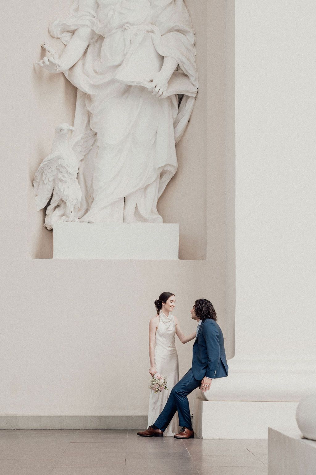 Wedding couple holds hands and gazes lovingly at each other beside a majestic white baroque sculpture.