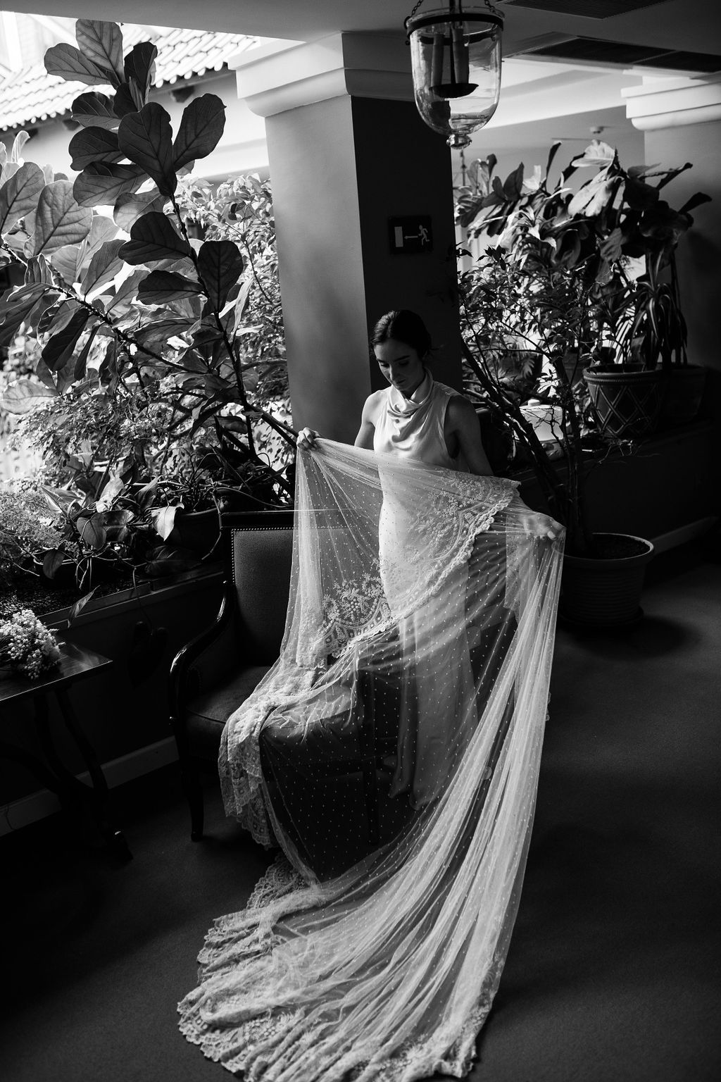 Bride in elegant lace wedding gown holds flowing veil among lush indoor plants in black and white photo.
