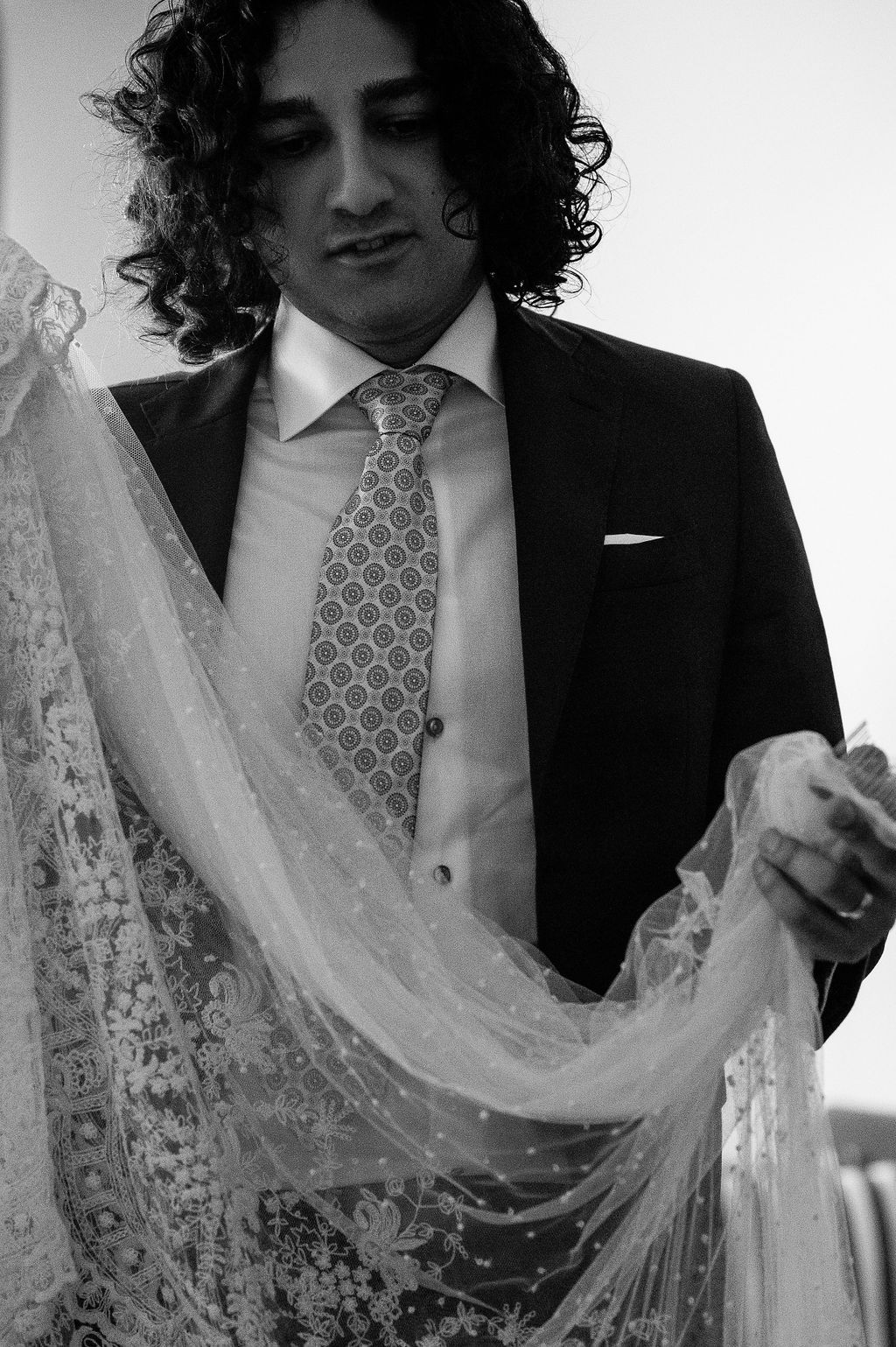 Black and white photo of a groom in a suit carefully holding delicate lace bridal fabric in his hands.