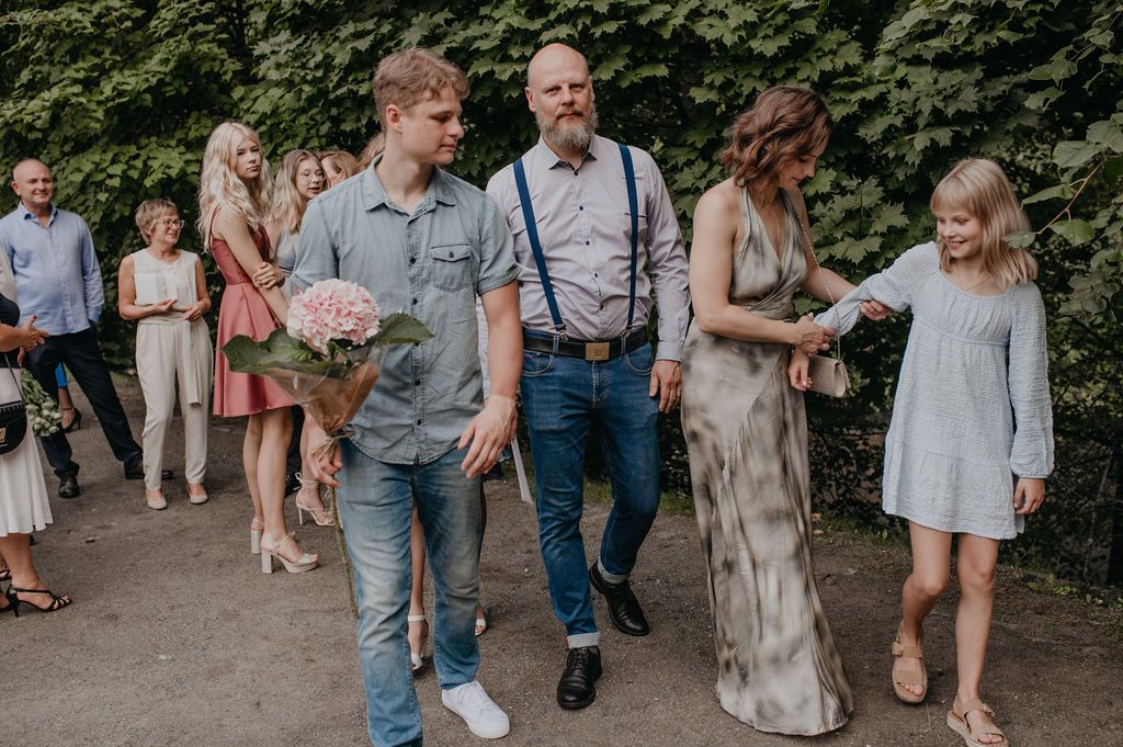 Wedding guests walk together outdoors, groom in suspenders holding bouquet of pink flowers.