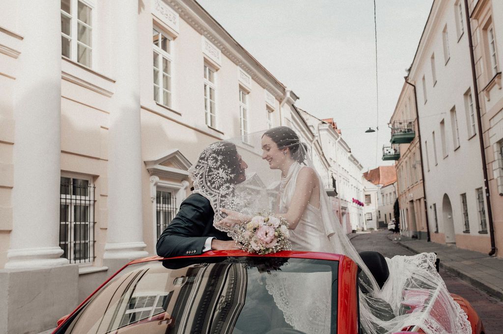 Bride and groom share a romantic moment standing through the roof of a red convertible on a city street.