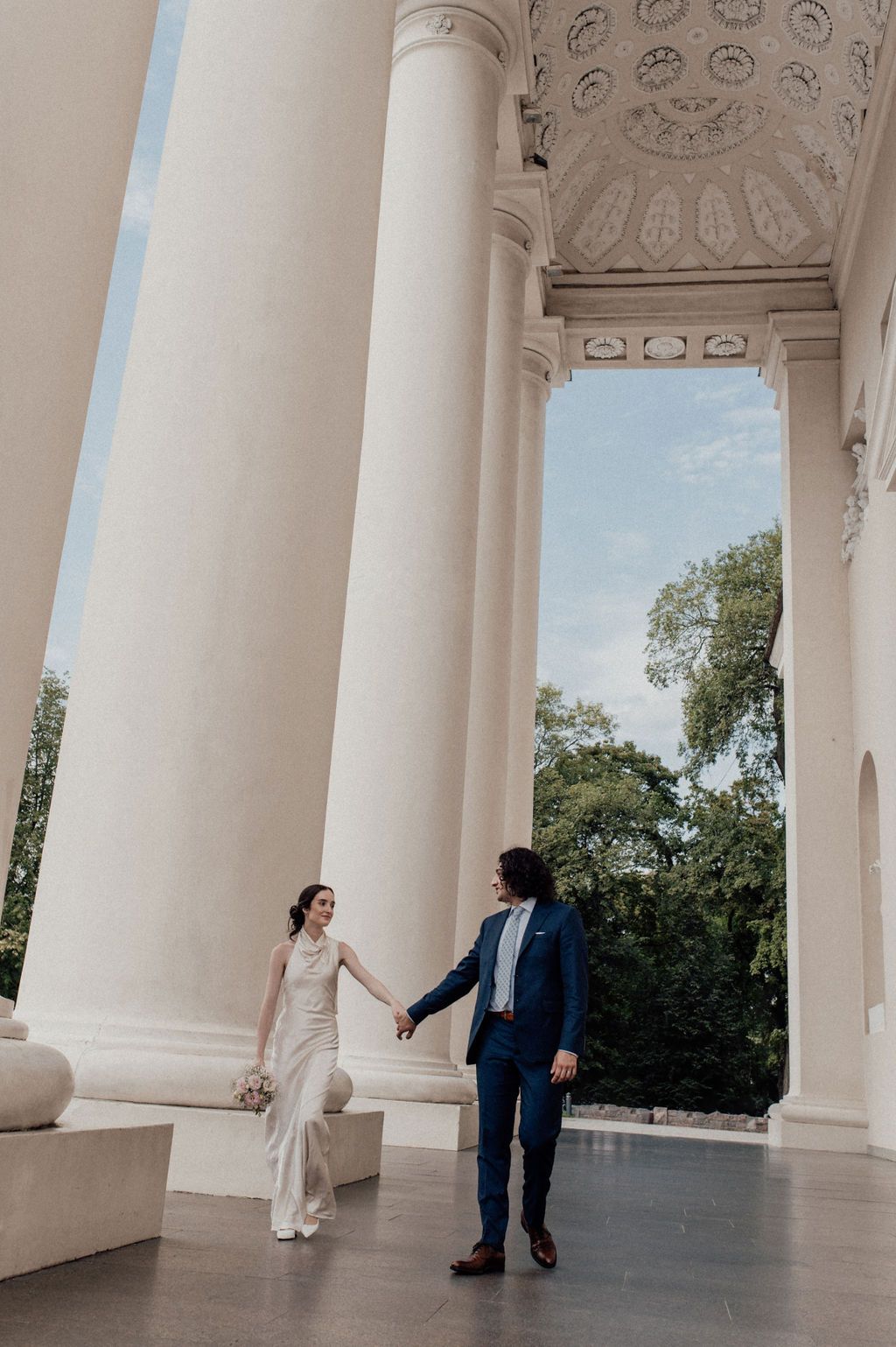 Bride and groom hold hands walking between grand white columns at an elegant outdoor wedding venue.