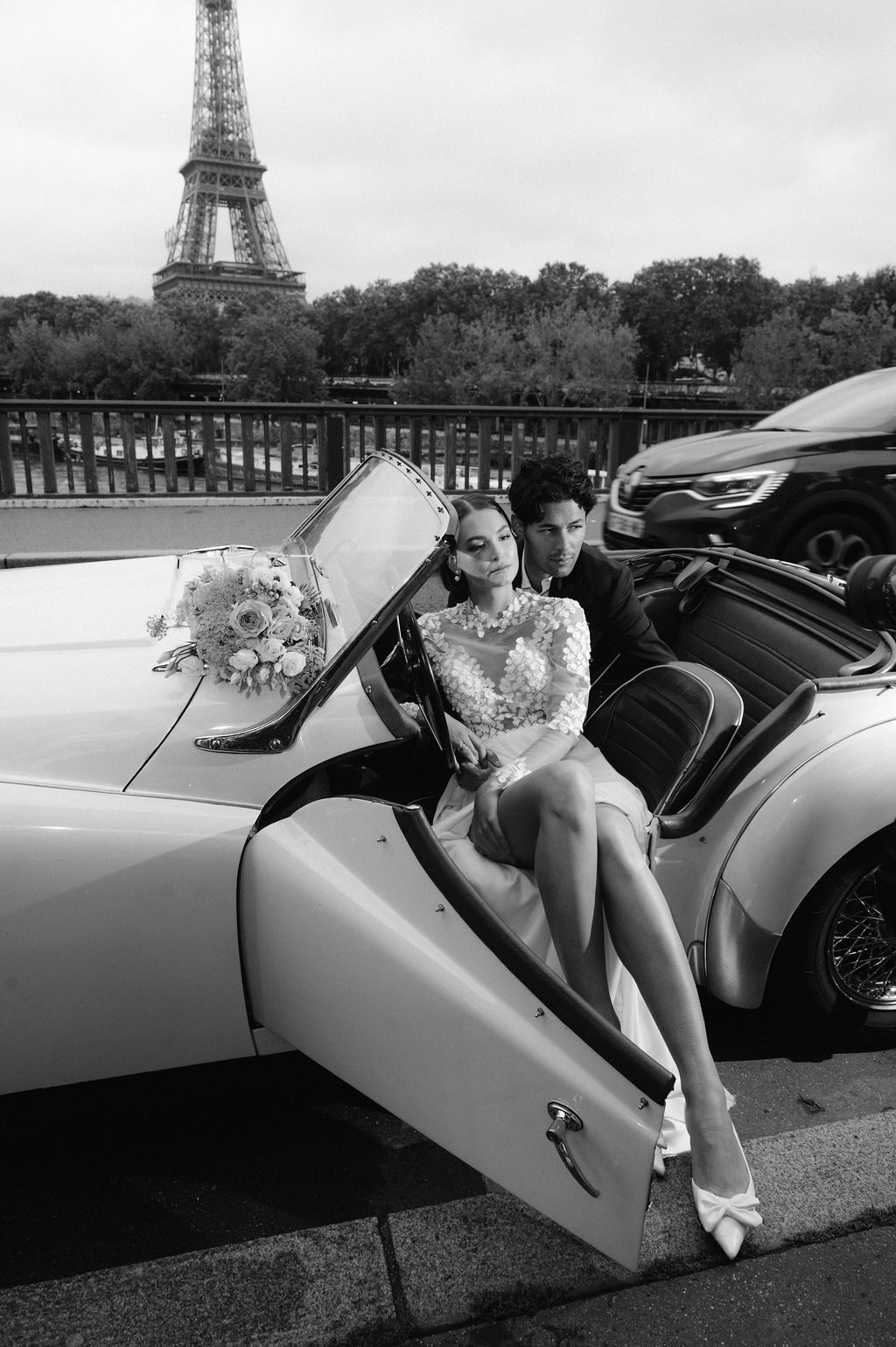 Bride and groom pose by a classic convertible car with the Eiffel Tower in Paris background.