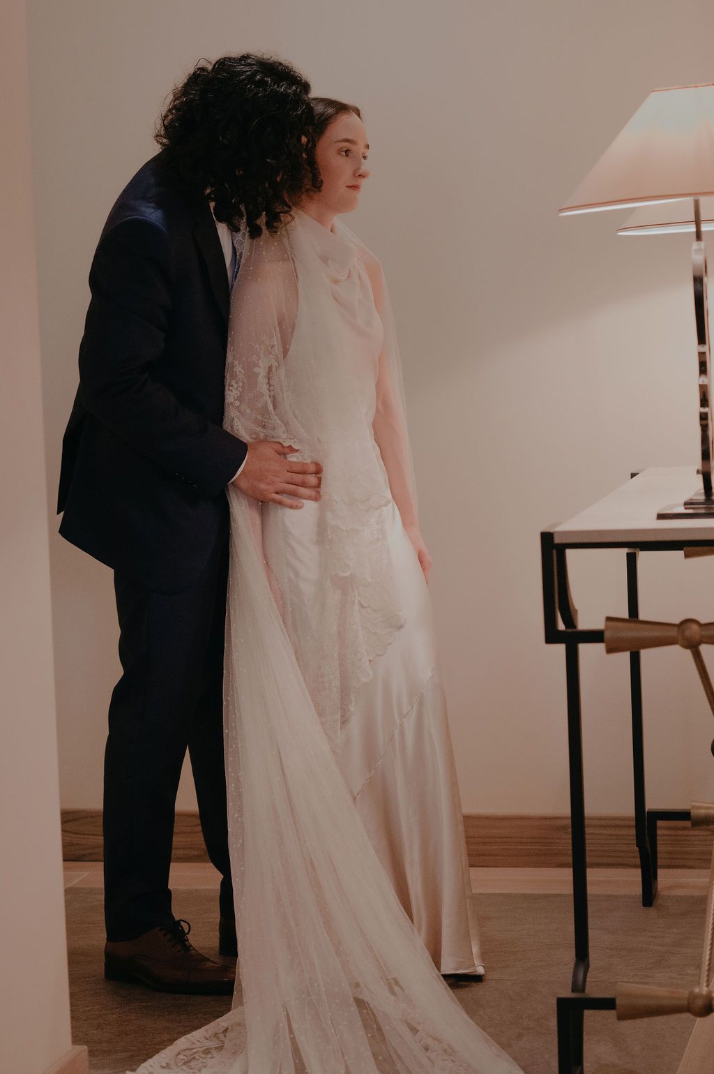 Bride in lace wedding gown stands with groom in navy suit in elegant indoor setting.
