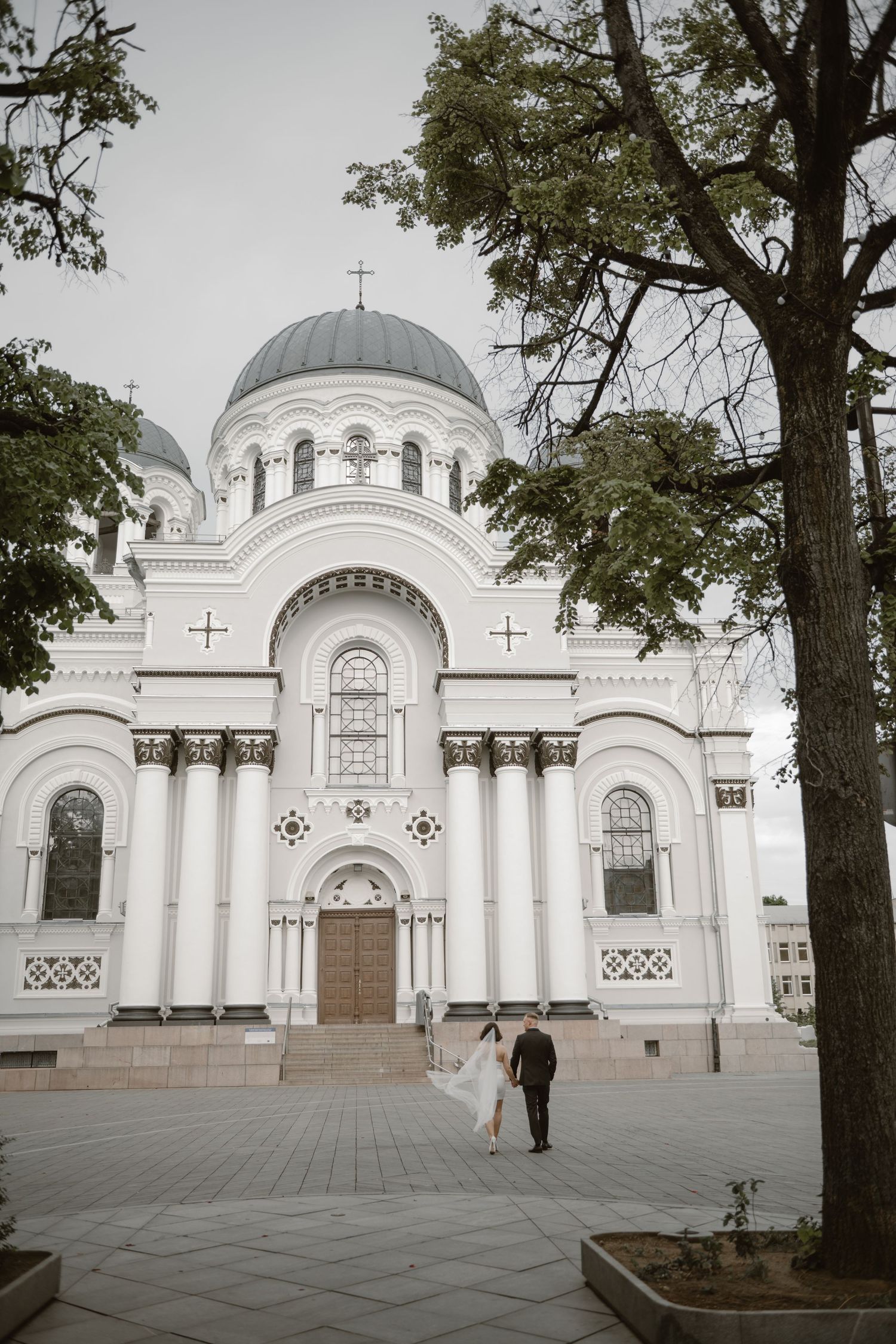 Bride and groom walk in front of a grand white Orthodox church with dome and cross on a cloudy day.