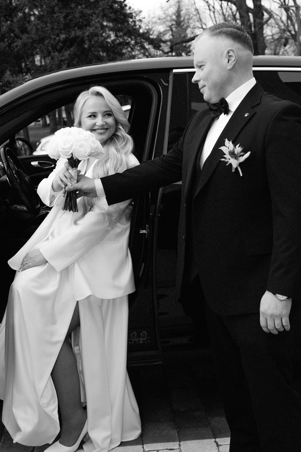 Black and white photo of a groom helping his bride out of a luxury car, bride holding white bouquet.
