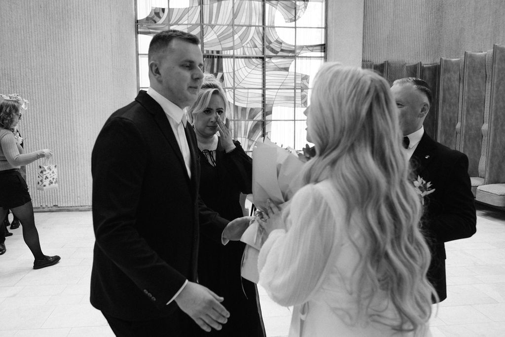 Black and white wedding photo of groom in suit greeting guests at elegant venue.