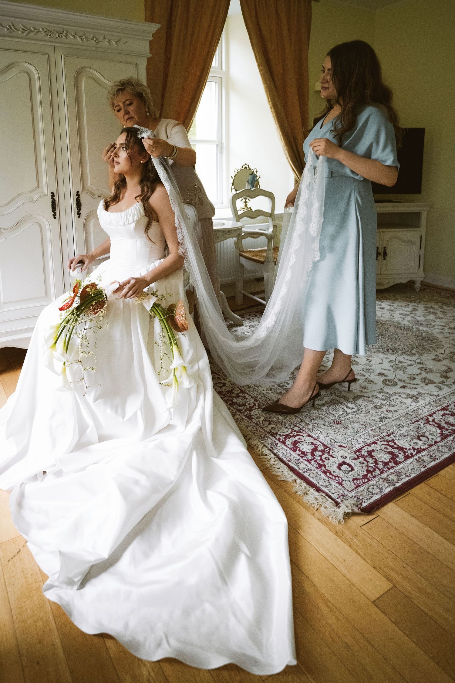 Bride in white gown holding bouquet as two women help arrange her veil in a classic elegant room.