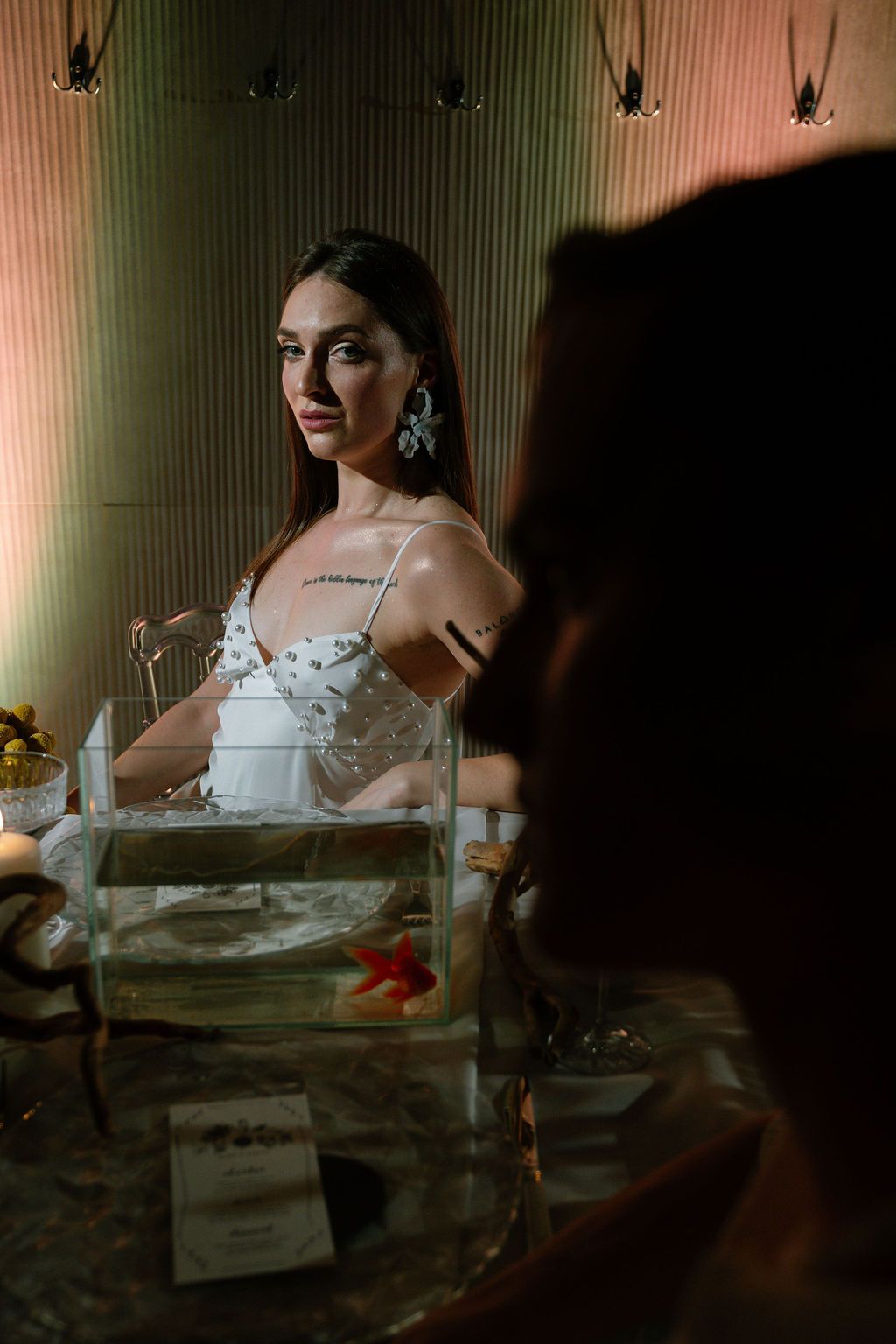 Woman in a white beaded dress and drop earrings sits behind a glass tank with a red starfish at an elegant candlelit table.