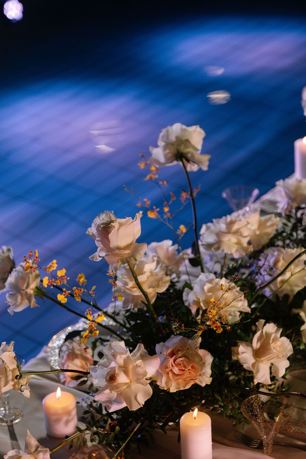 Cream and peach roses with orange accents form an elegant floral centerpiece beside lit candles on a table, with blue light on the floor.