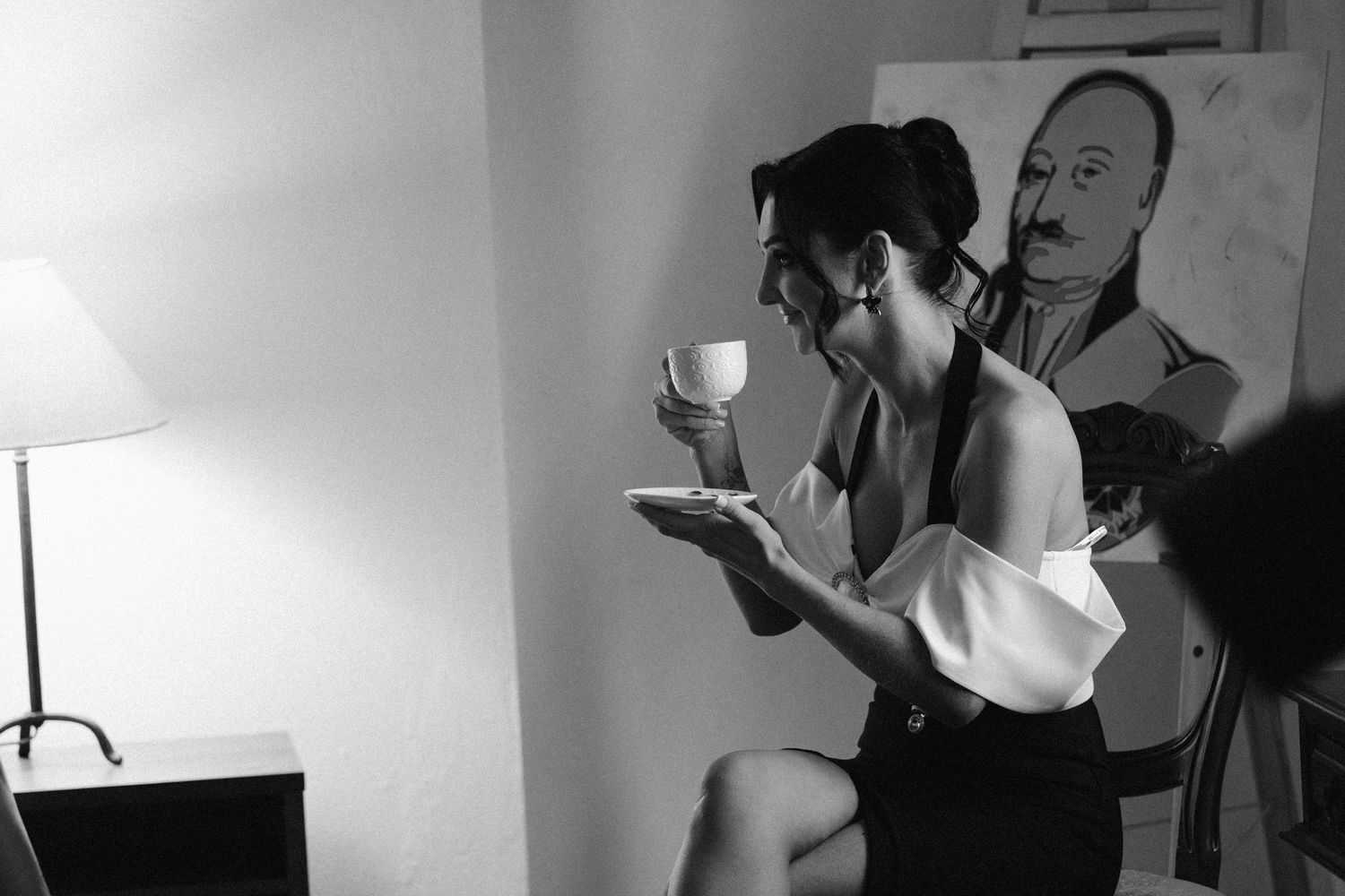 Black and white photo of a woman in an off-shoulder top sipping tea from a cup and saucer beside a lamp and a painted portrait.