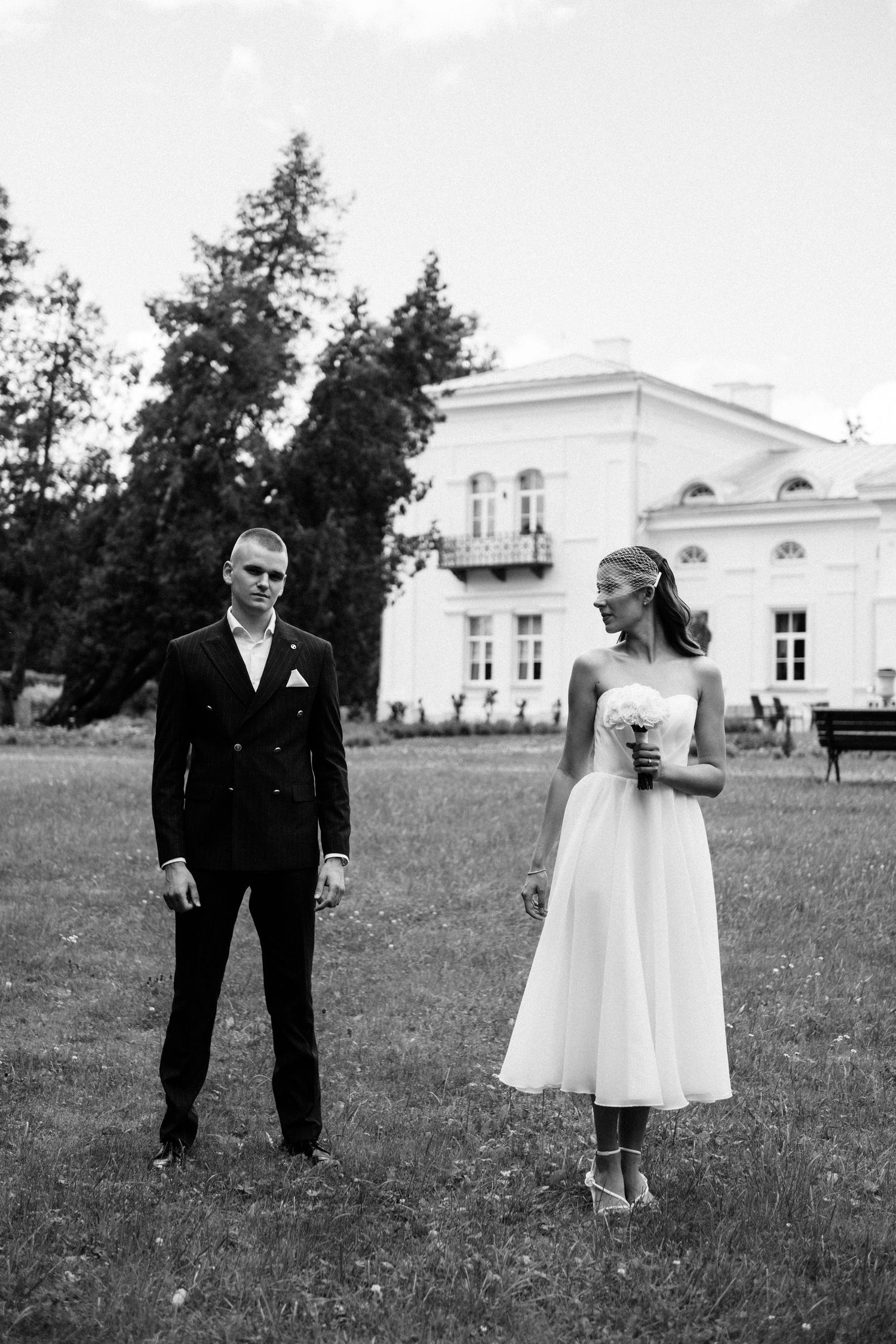 Black and white wedding photo of a groom in a double-breasted suit and a bride in a short dress holding flowers on a lawn.