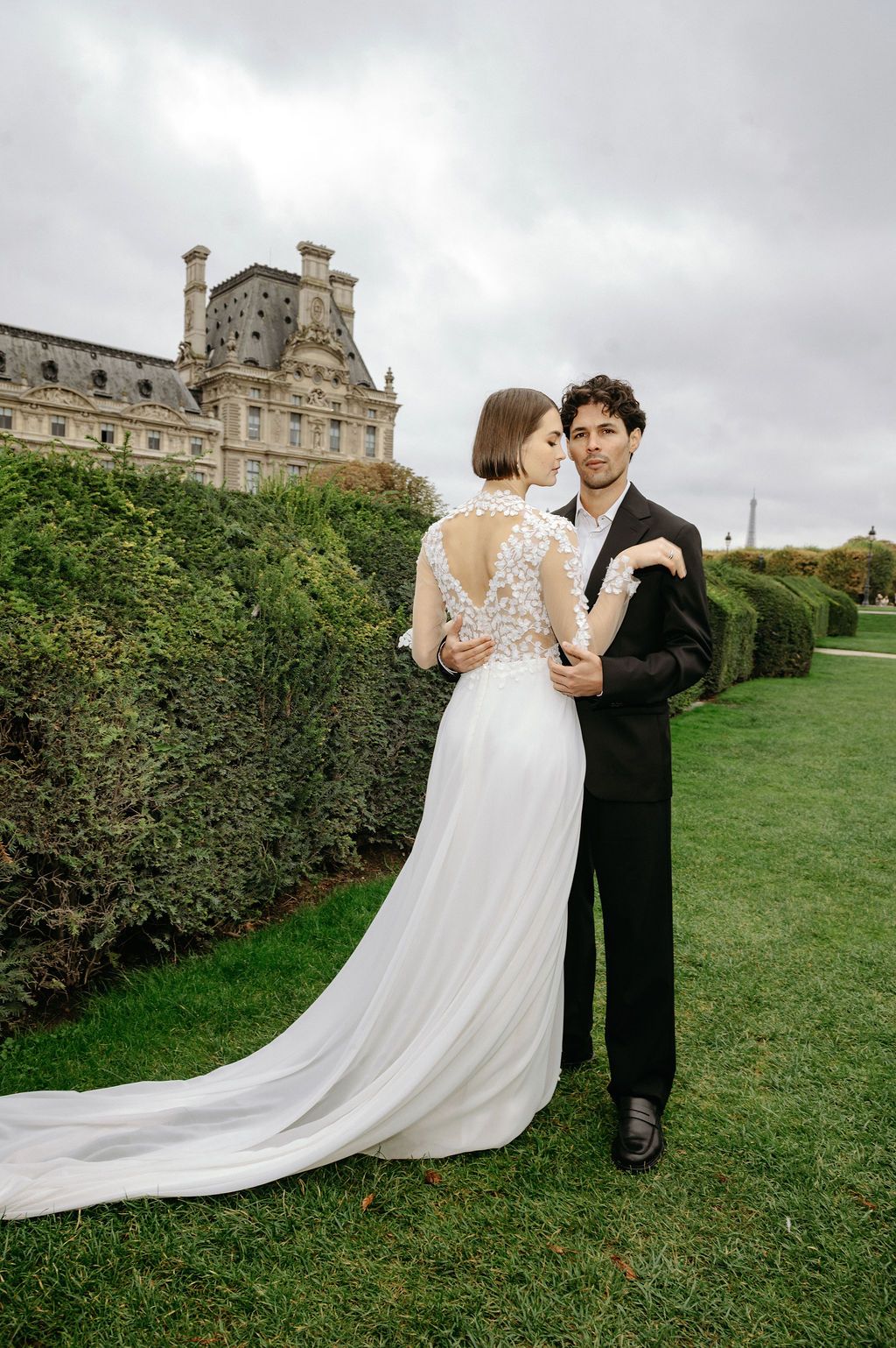 Bride and groom embrace in lush garden near the Louvre in Paris, bride wearing elegant lace wedding gown with long train.