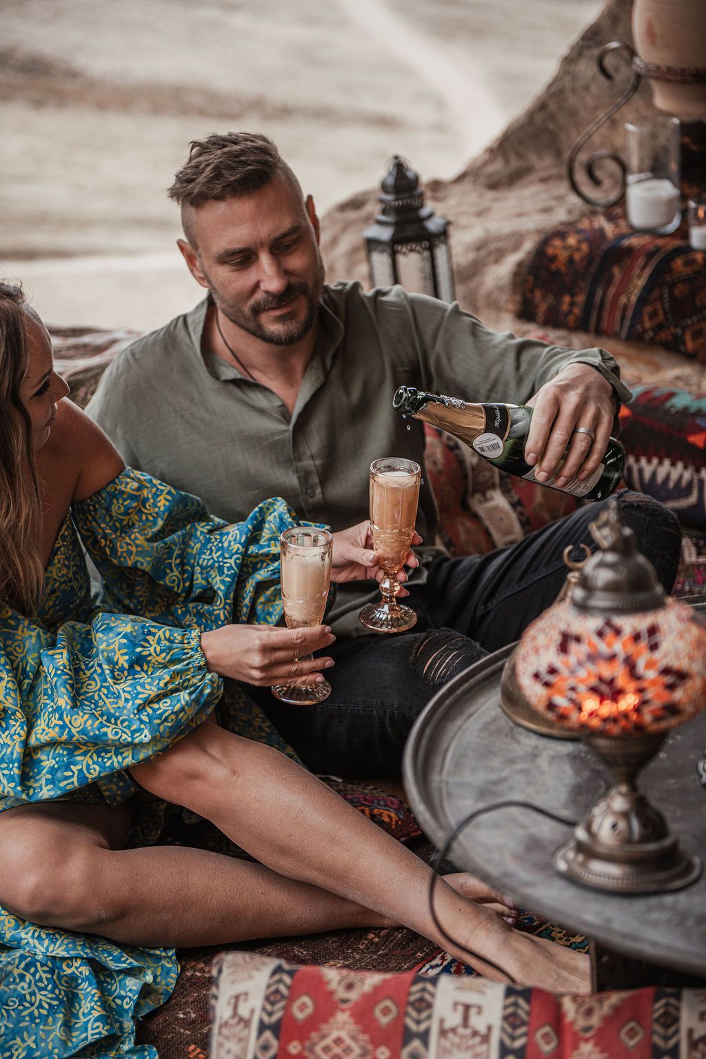 Couple sitting on vibrant Turkish rugs sharing champagne in a romantic desert boho picnic setting.