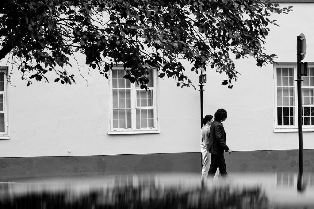 Black and white photo of a person walking past a white building with windows, under overhanging tree branches.