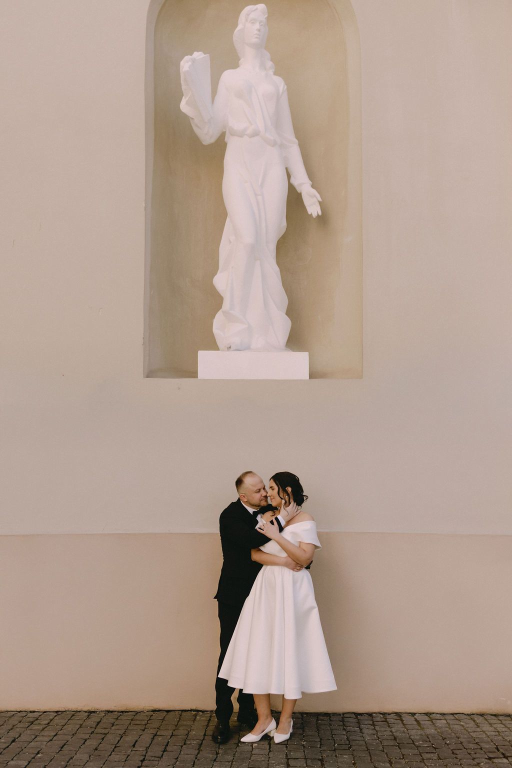 Bride and groom kissing beneath a white classical statue niche on a elegant building wall on their wedding day.