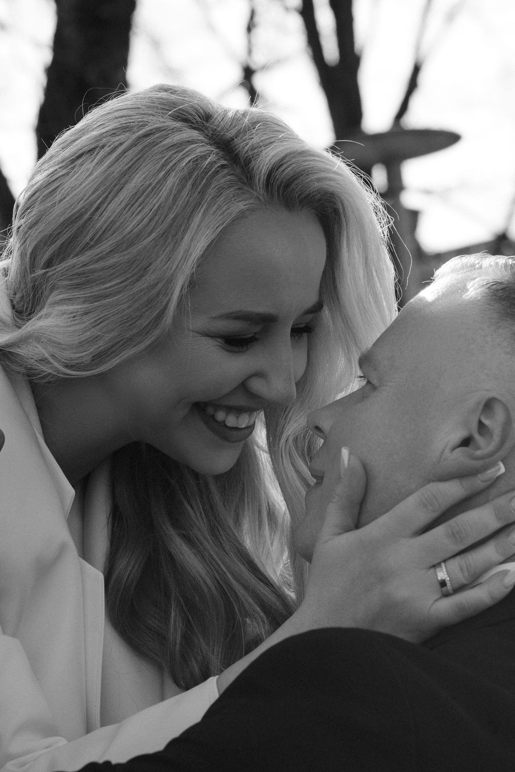 Black and white photo of a smiling blonde woman leaning in close to a man wearing a wedding ring.
