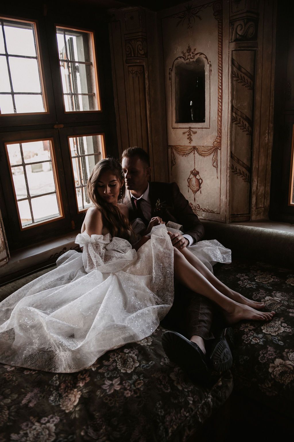 A bride and groom sit intimately together on a vintage sofa in an ornate room, her white lace gown draped elegantly around them.