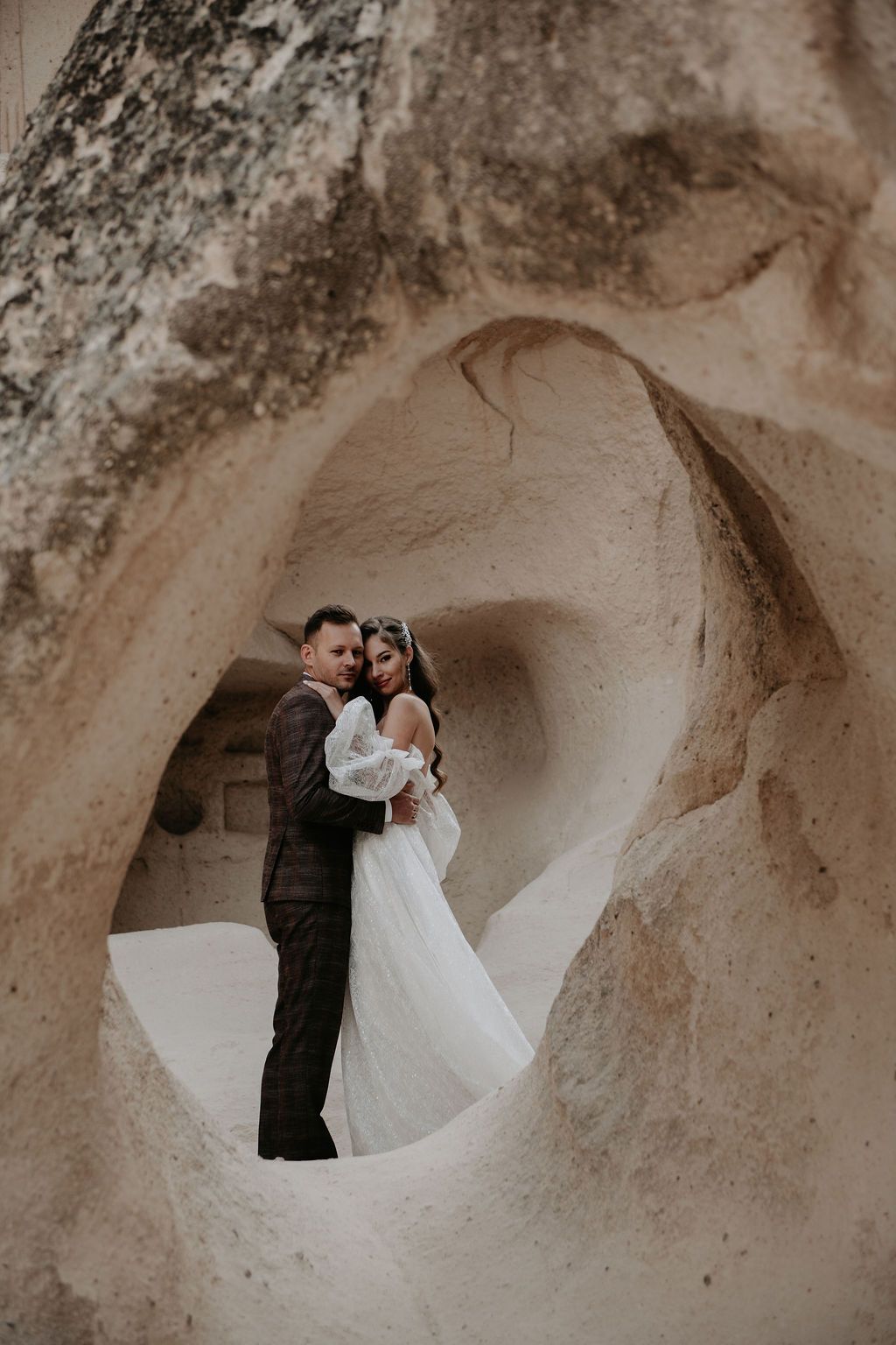 Couple in wedding attire embracing inside ancient cave rock formations in Cappadocia, Turkey.