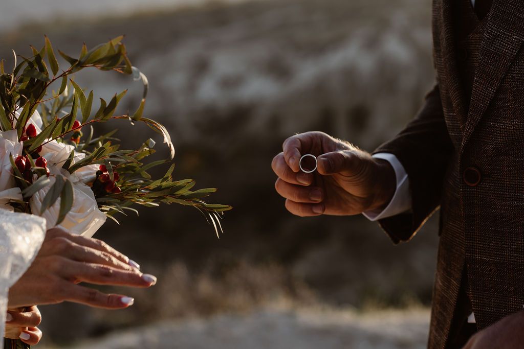 Groom holding a wedding ring while bride holds a floral bouquet with olive branches during an outdoor ceremony.