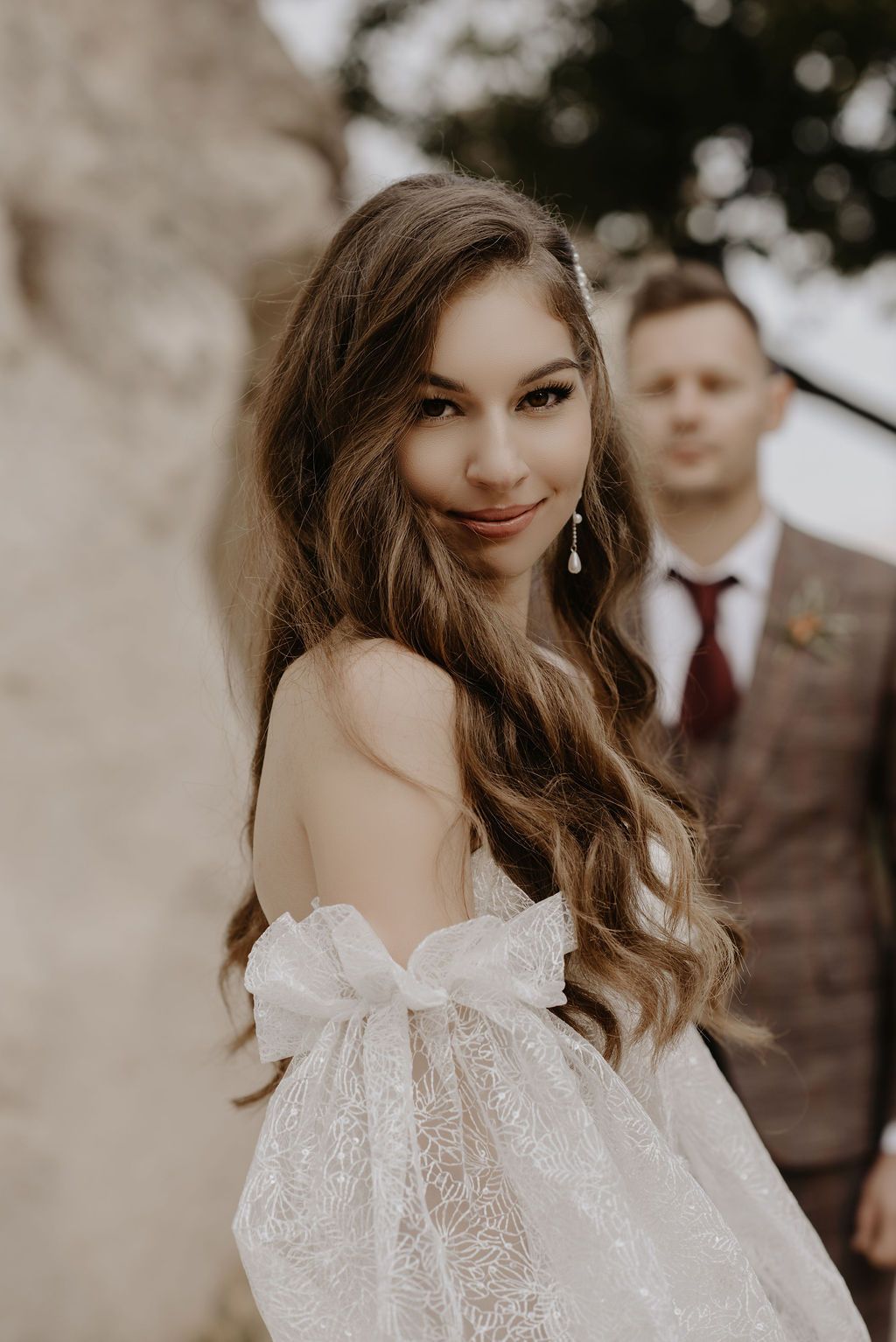 Bride in off-shoulder lace wedding gown with bow, long wavy hair, pearl earrings, groom in plaid suit blurred behind.