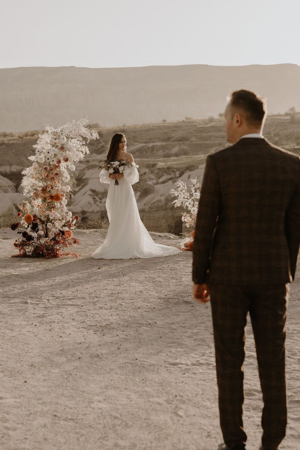 Bride in off-shoulder gown stands by floral arch in desert landscape as groom approaches.